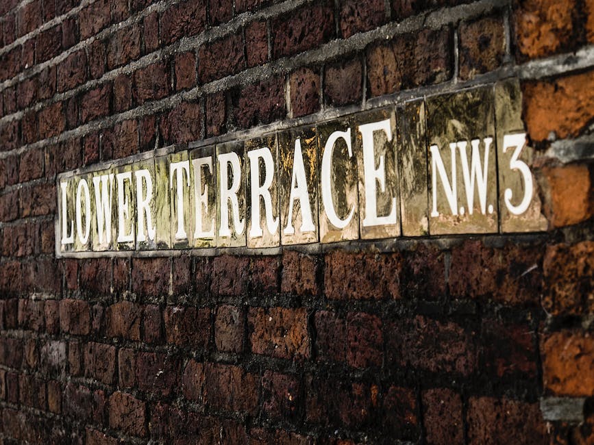 A close-up of a rectangular street sign mounted on a weathered red brick wall, displaying the text 'Lower Terrace NW3' in white capital letters on a reflective metal background. The sign appears aged with some dirt and moss around its edges, indicating outdoor exposure. The wall behind the sign comprises uneven bricks with dark mortar, typical of historic London buildings. This scene is relevant to house removals and relocation services, illustrating the type of address signage encountered during a home relocation or furniture transport process handled by Man and a Van Belsize Park, situated in NW3, near Haverstock Hill. The image captures the initial stages of moving logistics, where identifying the property at the street level is essential for planning efficient loading and unloading, often involving careful handling of furniture, boxes, and packaging materials near the building's entrance or pavement area.