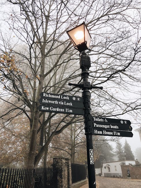 A black metal street lamp with a illuminated glass-enclosed light fixture on top, situated next to a leafless tree with numerous thin branches extending outward. Attached to the lamp post are multiple black direction signs with white text indicating nearby locations, including 'Richmond Lock', 'Isleworth via Lock', 'Kew Gardens 1/4m', 'Toilets & Passenger boats', and 'Ham House 1/4m'. The signs suggest a rural or park area within a historic or scenic part of Belsize Park, NW3, close to Haverstock Hill. In the background, there is a brick wall, a fence, and a few modest buildings or structures, with a slightly foggy atmosphere. This setting is relevant for home relocation or moving services, highlighting the local environment where furniture transport and packing might occur during a house removal.