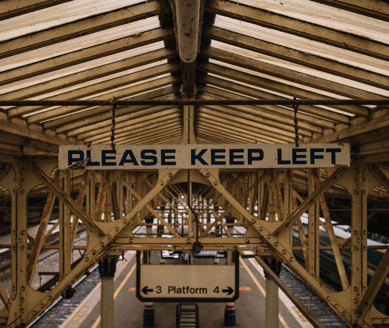 A large, rectangular railway station sign displaying directions and information for Ealing Broadway station, situated on the platform with a clear blue sky and green trees in the background. The sign